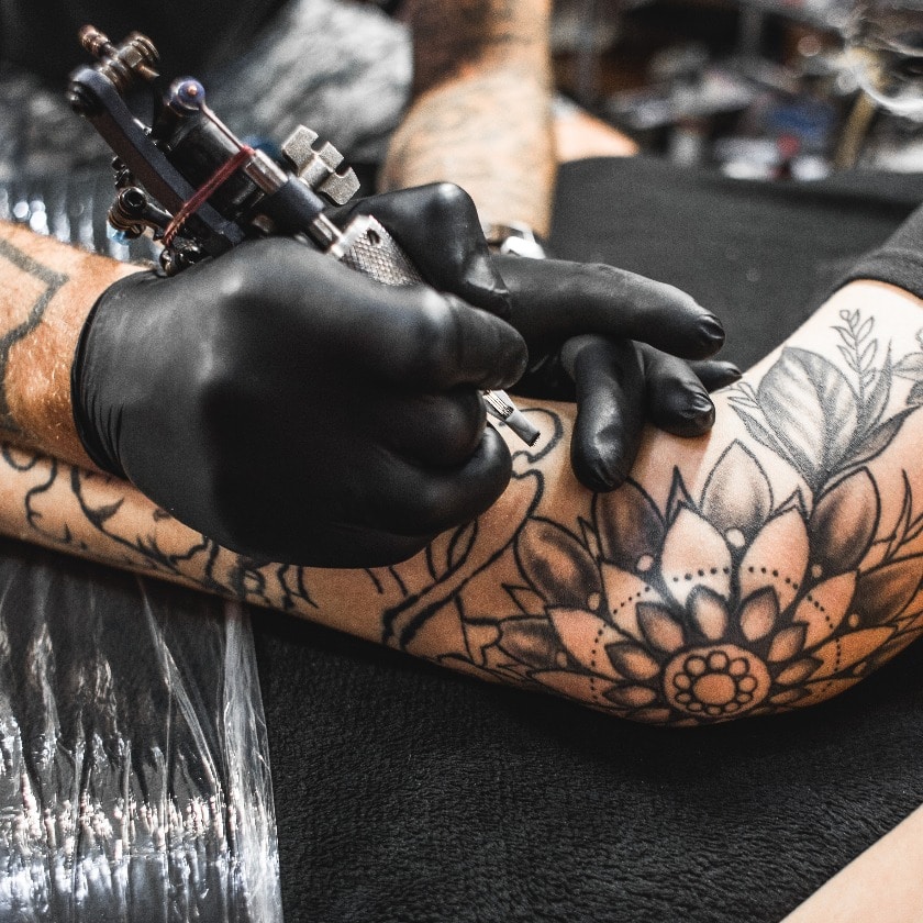 Mädchen mit Dreadlocks in einem Tattoo-Studio. Der Meister erstellt ein Bild auf dem Körper eines jungen, schönen Mädchens. Nahaufnahme der Hände und der Tätowiermaschine